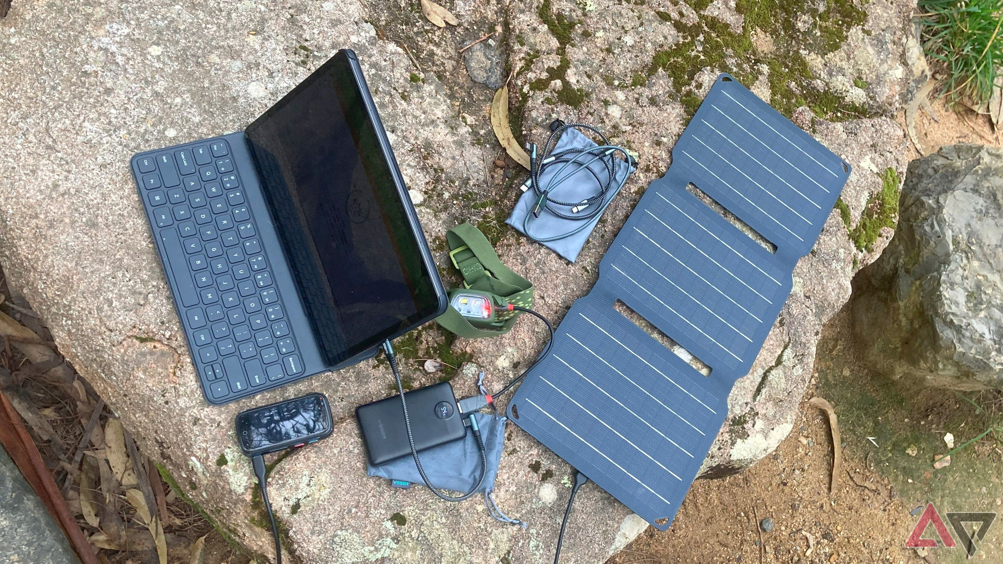 Carregador, banco de energia, painel solar, smartphone, farol e vários cabos em uma mesa de pedra na natureza